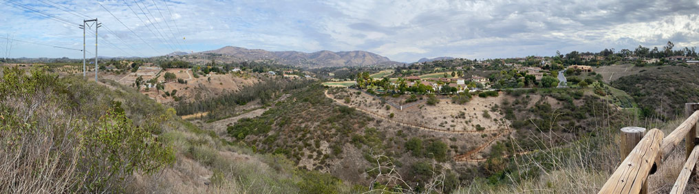 A panoramic hilltop view overlooking the green Santa Fe Valley and neighborhoods of Rancho Santa Fe with the mountain front of the Del Dios Highlands in the distance, Powerlines are on the left crossing the valley.