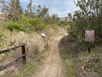 View of an earthen trail with a fence and small display on one side and a brown trail sign on the other.