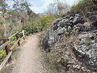 View of the earthen trail with a fence on one side and a dark rock outcrop on the other.