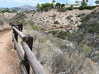 View of a fence rail along an earthen trail with switchbacks on a gray hillside in the distance.