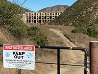 View of a gate with a Keep Out sign with Lake Hodges Dam in the distance beyond a trail.