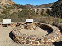 A low circular stone wall with display signs with Lake Hodges Dam in the distance.