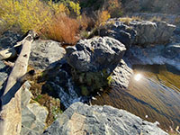 Large basalt outcrops and white granite boulders next to a waterfall and pool pool in the river bed.