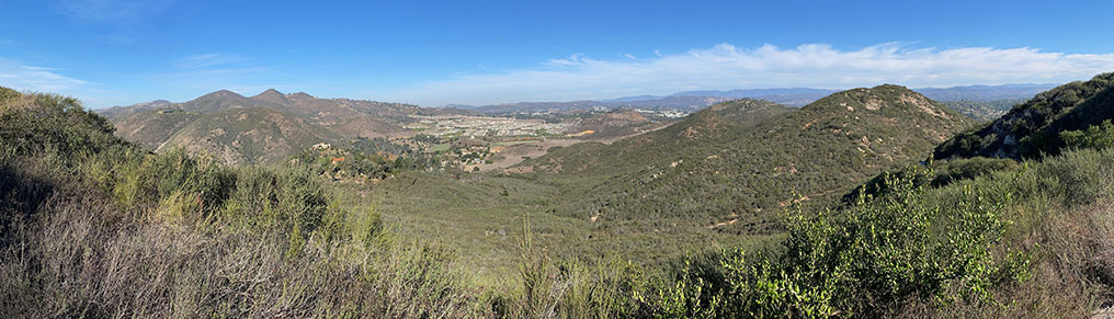 a Wide angle view of a broad, gently U-shaped valley with mountains covered with chaparral, with the Harmony Grove development in the distance. Mountain peaks highlight the highlands areas.
