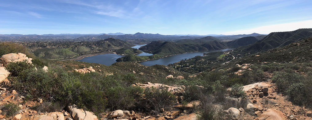 Wade-angle view of the Lake Hodges park area as seen from the Lake Hodges overlook. Rocky, chaparral covered mountain slopes are in the foreground. Distan peaks including the Cuyamaca mountains, Woodson Mountain, and Black Mountain are in the distance.