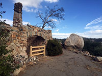 A stone arch and chimney, a short fence, and large boulder next to a drive next to the ruins of a burned out ranch house.