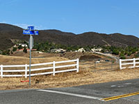Road interesction sing at Hayes Avenue and De Luz Road, with white board fence, rolling pature hils, ranch homes, and a dark mountain ridge in the distance of the Santa Rosa Plateau.