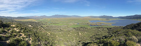 Panoramic view of Warner Valley view from the Henshaw Scenic Vista Observation Site. The Elsinore Fault Zone is beyond Lake Henshaw running along the linear mountain front of Mesa Grande. The linear stream canyon in the foreground follows one of several faults that parallel the trend of the Elsinore Fault into the lake area. The small dark area among the green grassy area is fault-bounded Monkey Hill. The yellow areas are field of California poppies blooming in the Warner Ranch area.