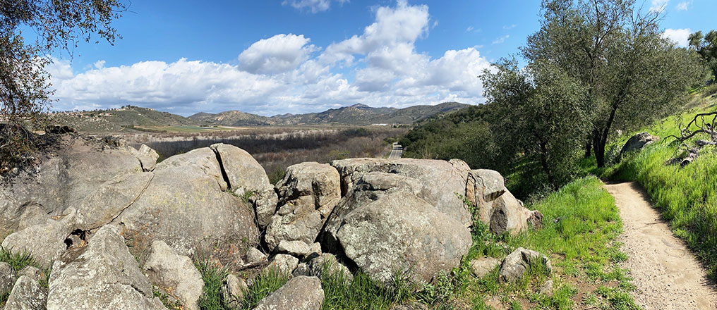 A large granite outcrop and boulders along the dirt trail. One of the boulders has a flat top, and Highland Valley Road appers above it. The rural San Pasqual Valley and surrounding mountainous ridges are in the distance.