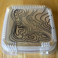 Countour lines illustrated by lines drawn on a stack of clear plastic box lids. Each lid represents a seperate elevation.