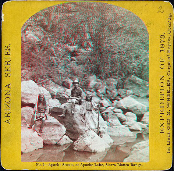 Two men (indians) with guns, and a third white man, sitting on boulders along the shore of a river.