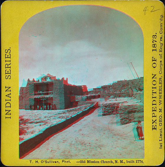 A rugged adobe chuch building surrounded by walls and adobe residences. People are standing in front of the mission.