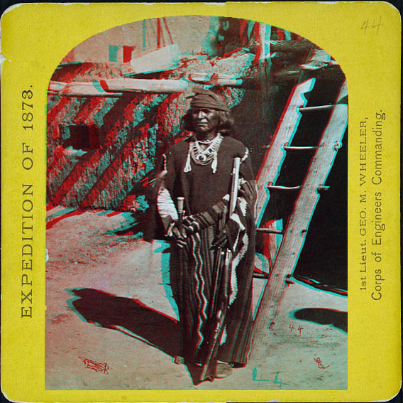 Man in full indian dress with gun and sword standing next to a ladder leaning against an adobe home.