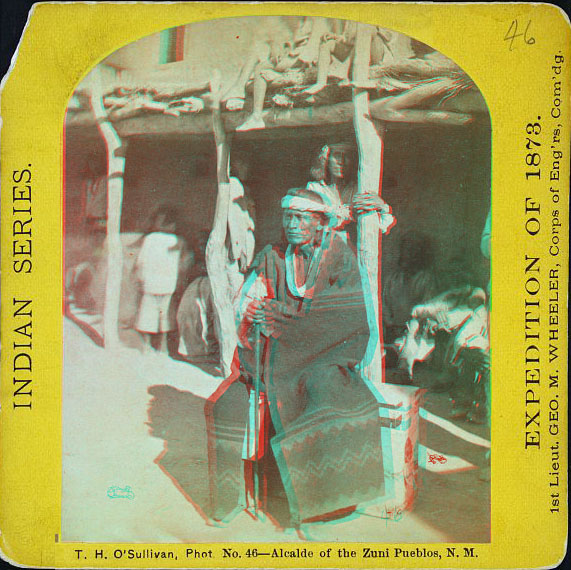 Man in full indian dress with a gun sitting in front of an adobe building with other people in the shade of an overhang.