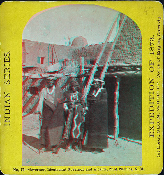 Three indians in native dress standing in fron of a ladder in front of an adobe building.