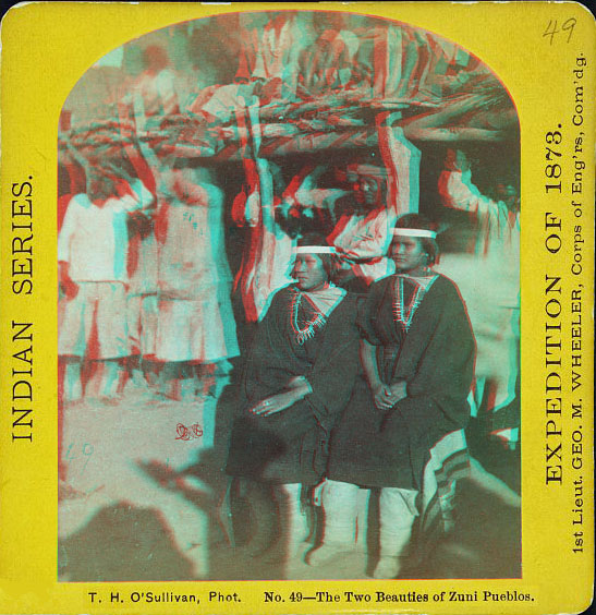 Two women in full native dress sitting with other people standing in the background in front of an adobe building