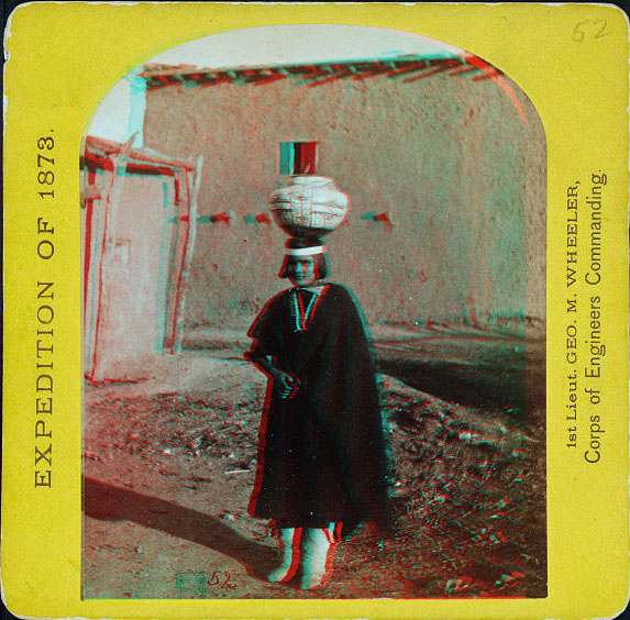 A woman in a dark dress with a large pot balanced on her head in front of adobe buildings