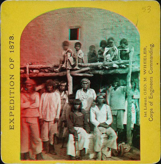 A group of men and children sitting and standing in front of an adobe building. Some are sitting on the roof of a pateo overhang.