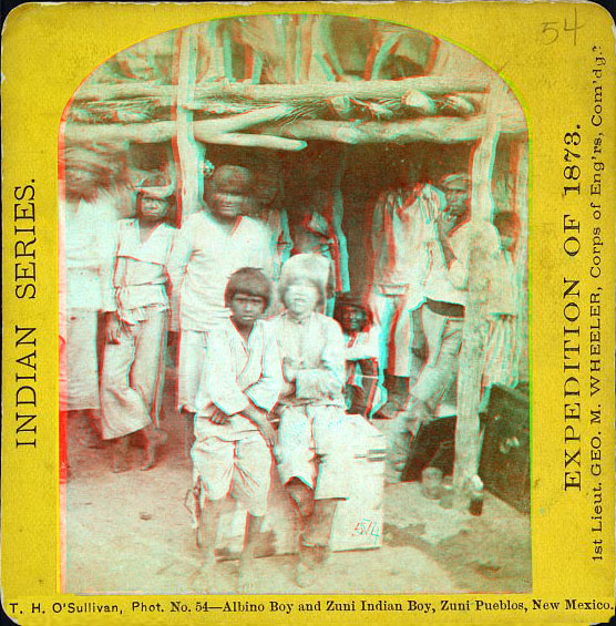 A group of indians sitting or standing in front of an adobe building. One child in front is an albino.