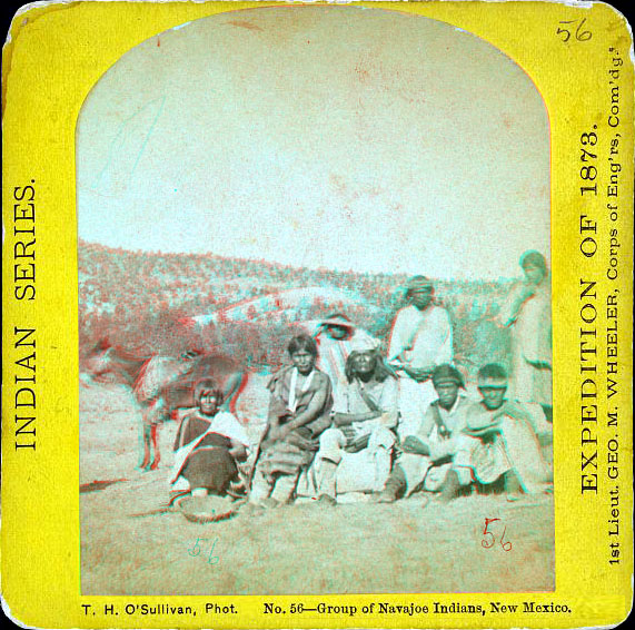 A group of people sitting or standing wearing native dress. A saddled horse is on the left, and a forested hill is in the background.