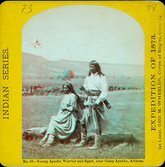 A woman sitting, and a man standing, wearing native dress. A hill is in the distance across a grassy plain.