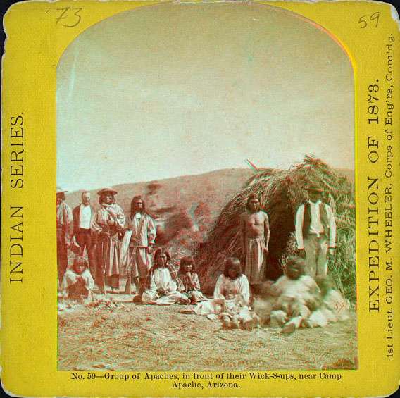 A group of people in native dress sitting or standing, one on horseback, next to a low round home made of sticks.