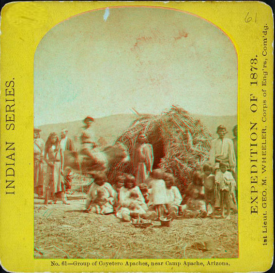 A group of people in native dress sitting or standing, one on horseback, next to a low round home made of sticks.