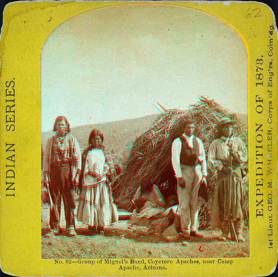 A group of people in native dress sitting or standing, one on horseback, next to a low round home made of sticks.