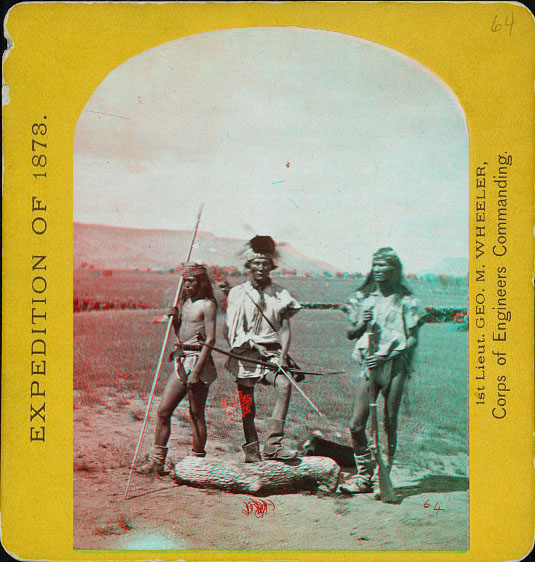 Thre men posed in native dress. One has a spear, one has a bow and arrow, another is holding a rifle.