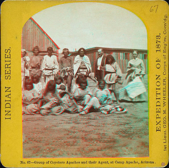 A group of posed people sitting or standing in front of some anglo-style buildings. One man and a woman in the group are anglo, the rest are natives.