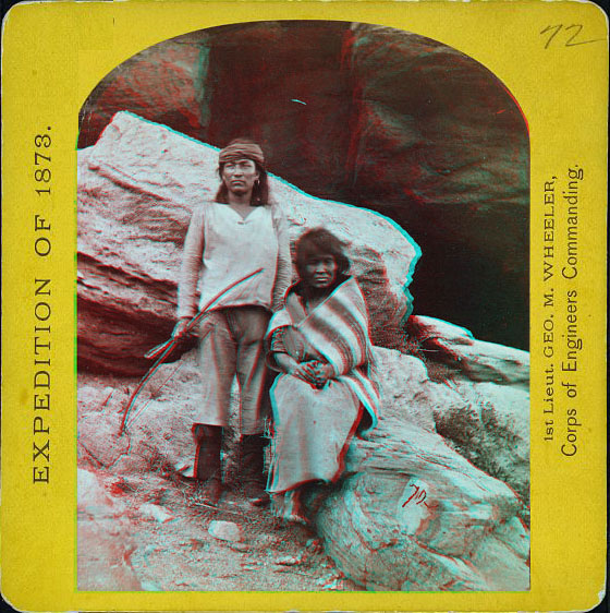 A man standing with a bow and arrow, and a woman sitting, in native dress posed in front of some large rocks.