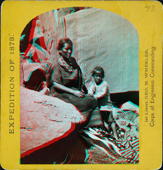 A woman sitting with a child next to her standing on a blanket. Large rocks are exposed in front and behind them.