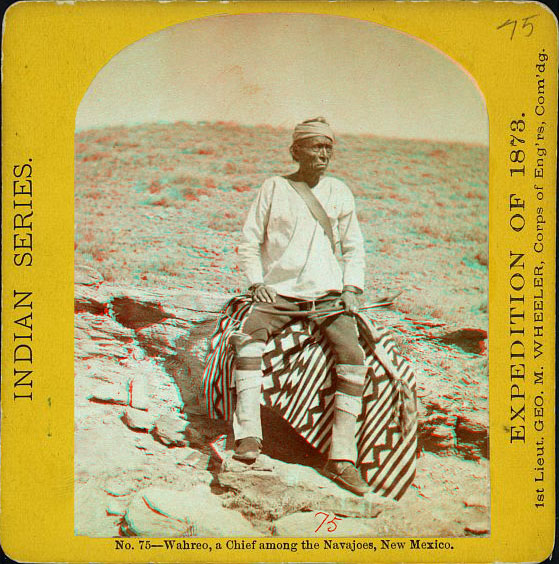 A man in native dress with a bow, sitting on a blanket on a rock.