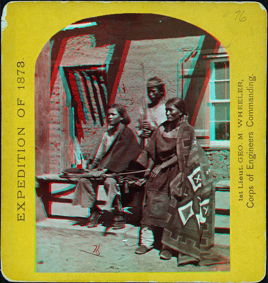 Three young people in native dress, in front of a window of an adobe building.