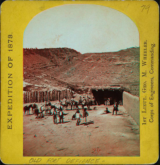 People standing and on horses in a barren outdoor corral surrounded by high fences, and a barren hill in the distance.