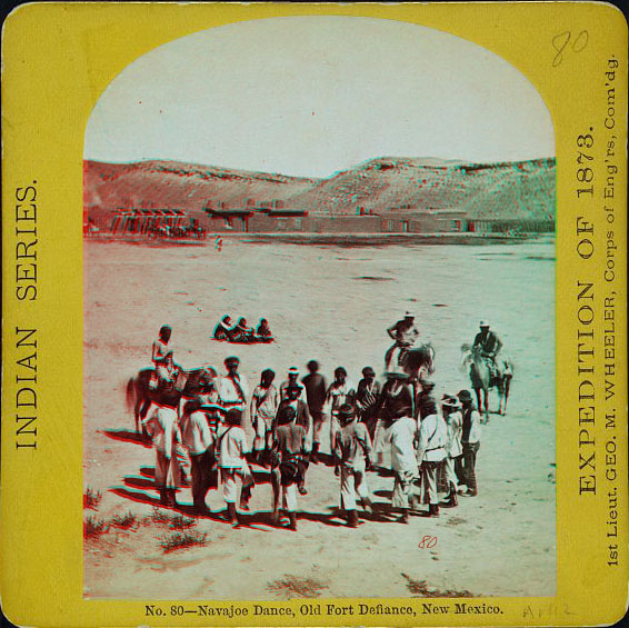 A circle of people in native dress, some holding legs in the air, three people on horseback, and three people sitting. A low adobe fort and hill are in the distance.