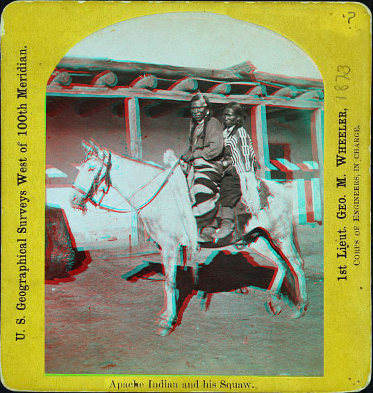 A man and a woman, in native dress, sitting on a horse in front of a low building.