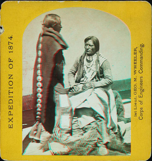 A man sitting, and a woman standing, facing him. Both are in native dress.