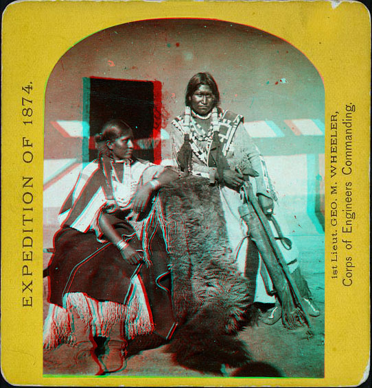 A man and a woman posed in front of a building with a buffalo skin hanging between them.