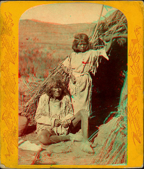 An old man sitting, and a child standing, in front of a dwelling made of sticks. 