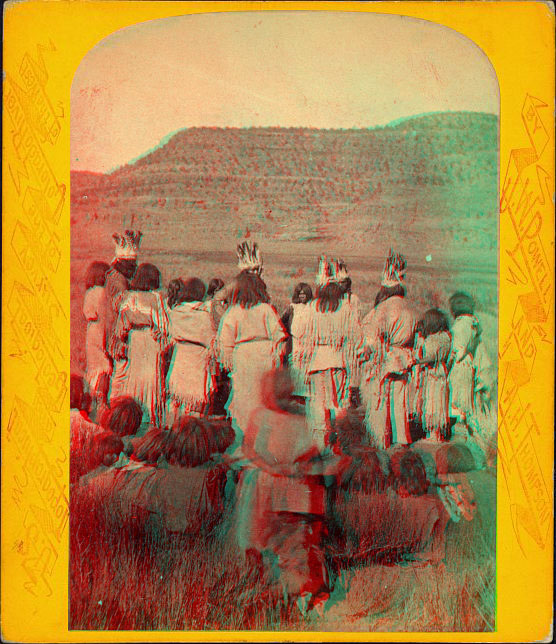 A group of people in light-colored native dress standing in a circle, some with crowns of feathers. Other people are sitting in the foreground with their backs toward the camera