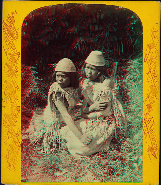 Two women in native dress, sitting, and wearing woven caps