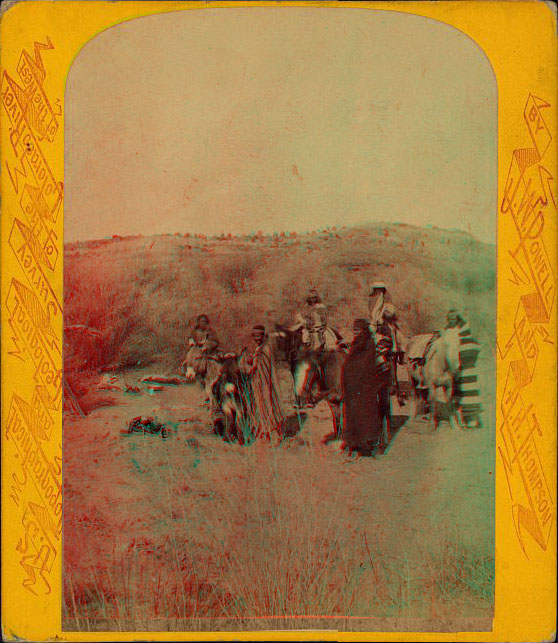 A group of people in tative dress sitting and standing, two are on horses. Another horse and a burrow are saddled