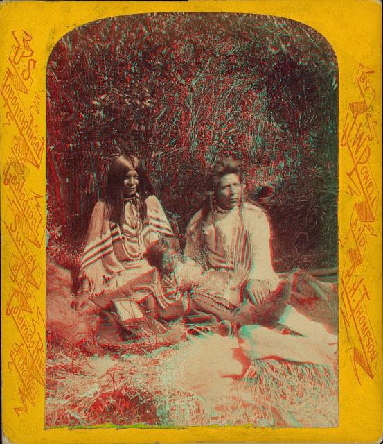 A man and a woman in full native dress, sitting outside on a blanket