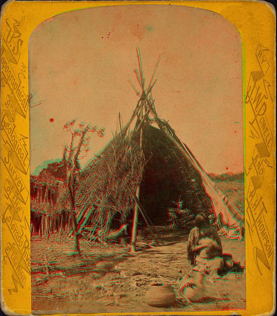 View of a wonan and a child sitting in fron of a dwelling made of sticks. Two clay pots are in the foreground