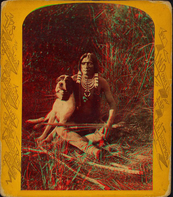 A young man sitting on the ground holding is dog and bow and arrows