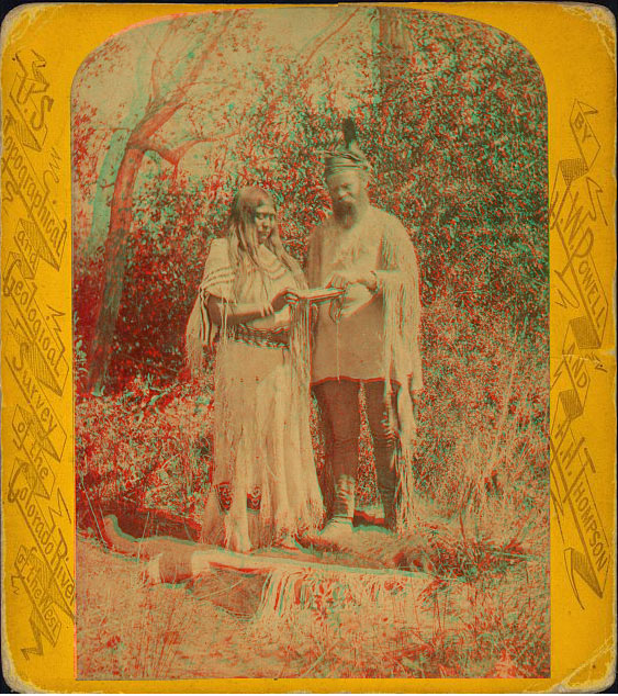 A woman in full dress looking at an object, standing next to a white man with a long beard.