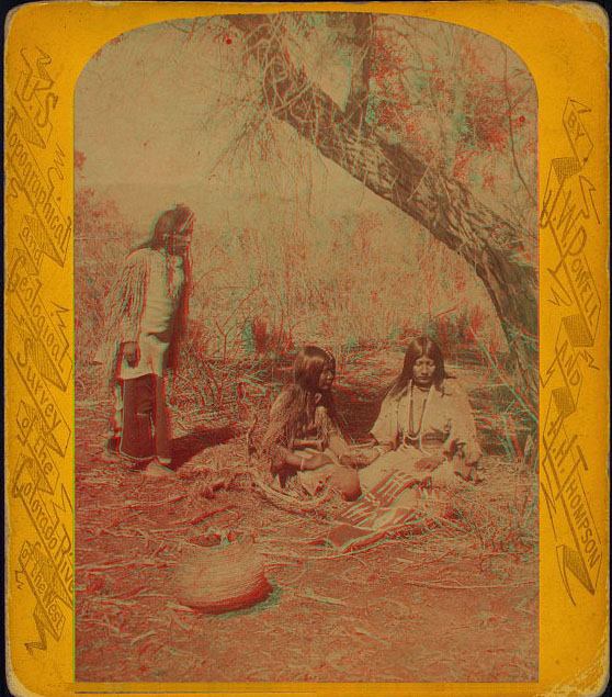 Two women in native dress sitting under a tree near a clay pot. An older man is standing nearby.